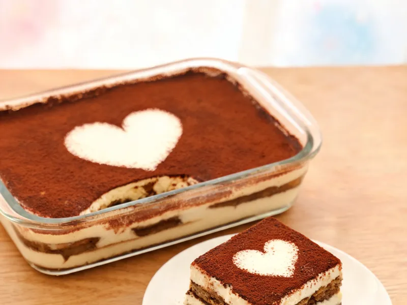 Tiramisu in einer Glasform, ein Stück auf dem Teller mit Kakao-Herz (KI-generiertes Bild)