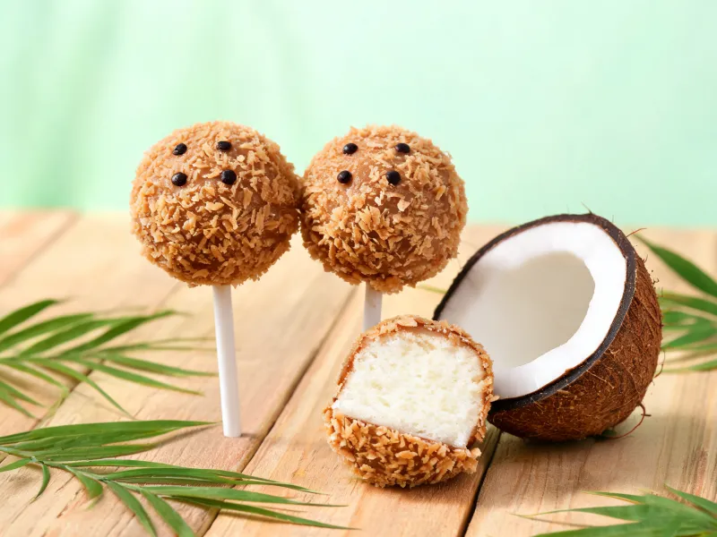 Cake Pops mit brauner Kokosraspel-Hülle wie Kokosnüsse, einer halbiert mit weißem Kokoskuchen innen (KI-generiertes Bild)