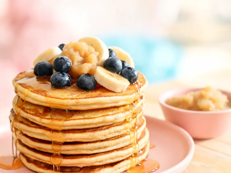 Stapel fluffiger Pancakes mit Apfelmus, Blaubeeren, Bananenscheiben und Ahornsirup (KI-generiertes Bild)