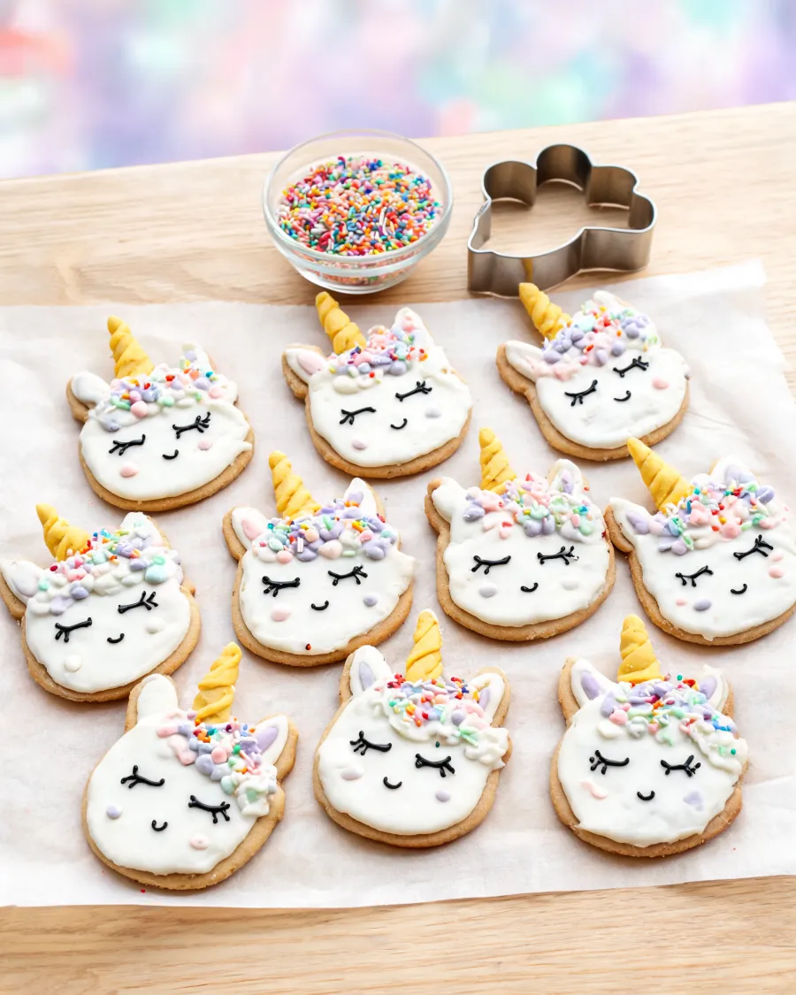 Ausgestochene Butterkekse als Einhornköpfe mit weißem Zuckerguss, goldenem Horn und bunter Mähne (KI-generiertes Bild)