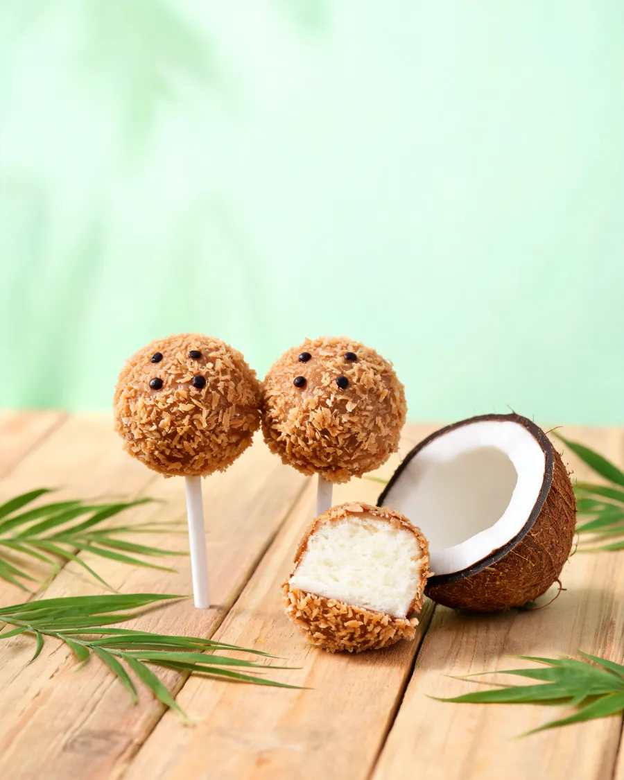 Cake Pops mit brauner Kokosraspel-Hülle wie Kokosnüsse, einer halbiert mit weißem Kokoskuchen innen (KI-generiertes Bild)