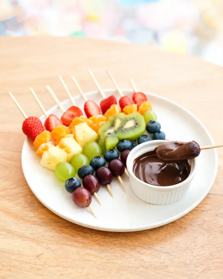Obstspieße in Regenbogenfarben mit Erdbeeren, Orange, Ananas, Trauben, Kiwi und Blaubeeren, daneben Schoko-Dip (KI-generiertes Bild)