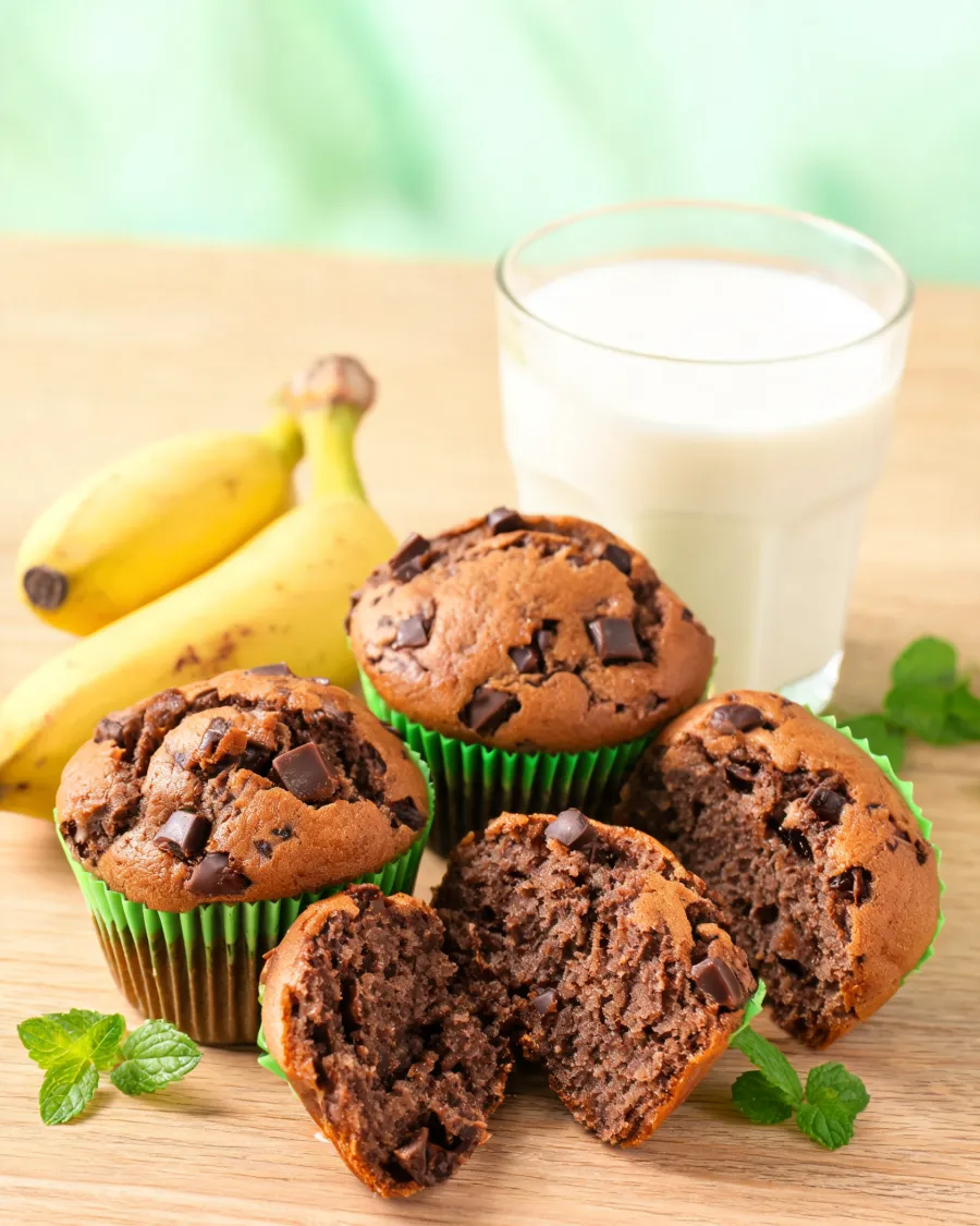 Schoko-Bananen-Muffins in grünen Förmchen, einer aufgebrochen, daneben Bananen und Haferdrink (KI-generiertes Bild)